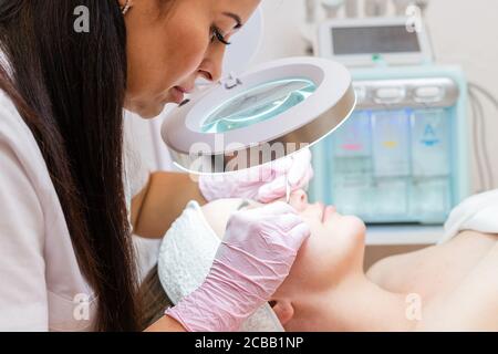 Il cosmetologo in guanti di gomma guarda attraverso una speciale lente d'ingrandimento e fa una pulizia meccanica del viso del cliente. Concetto di professio Foto Stock