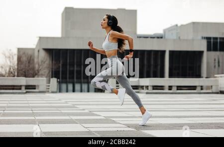 Foto a tutta lunghezza di una donna sportiva che si springe in città. Donna in abbigliamento sportivo e cuffie che corrono velocemente all'aperto. Foto Stock