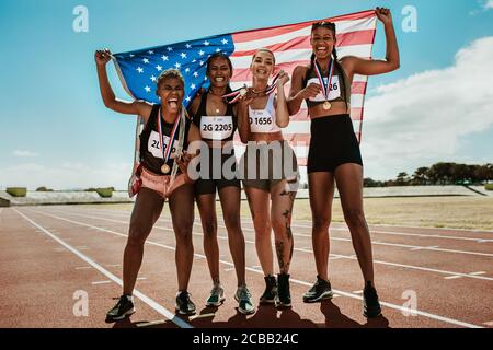 Felici atleti multirazziali che festeggiano la vittoria mentre si levano in piedi insieme in pista. Gruppo di corridori con medaglie e bandiera nazionale americana vincendo un Foto Stock