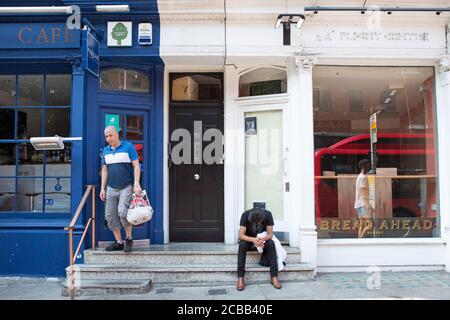 Un uomo si siede stanco fuori da un negozio che è chiuso in Hampstead High Street come affari vicino tutto il Regno Unito a causa del Coronavirus o Covid-19. Foto Stock