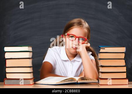 Ritorno a scuola. Studente di bambina seduto al tavolo sullo sfondo di una lavagna nera. Scolaretta stanca con mucchi di libri. Foto Stock