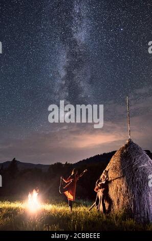 Una coppia romantica che si riposa in montagna, una ragazza che balla accanto al fuoco e un ragazzo che suona la chitarra si appoggiò su rick di fieno secco sotto un incredibile cielo notturno pieno di stelle e Via Lattea Foto Stock