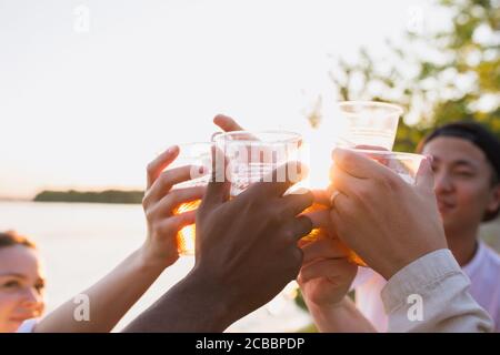 Primo piano. Gruppo di amici che indossano bicchieri da birra durante il picnic in spiaggia sotto il sole. Stile di vita, amicizia, divertimento, weekend e riposo concetto. Sembra allegro, felice, festoso, festoso. Foto Stock