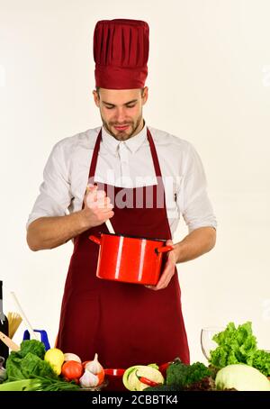Cucina lavori in cucina vicino a tavola con verdure e utensili. L'uomo in cappello  e grembiule si mescola in pentola. Concetto di preparazione degli alimenti. Chef  con viso eccitato tiene tegame rosso