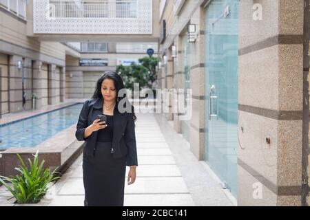 Una donna indiana in piedi guardando nel suo telefono in un ambiente urbano aziendale. Dipendente femminile aziendale in abbigliamento formale intelligente Foto Stock