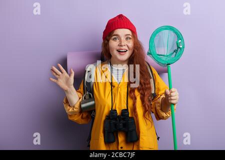 emozionato escursionista allegro indossare tappeto di cura, zaino, cappotto giallo, binocolo sul collo tenendo la rete farfalla, primo piano foto. isolato sfondo blu, st Foto Stock