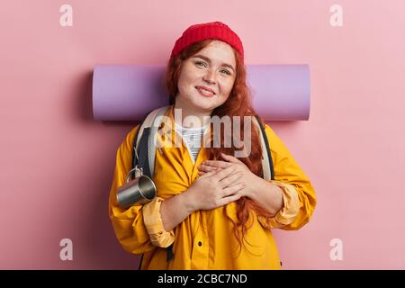 donna che indossa zaino, zaino, cappotto giallo, camicia verde e berretto in maglia rossa ha un'espressione amichevole, premendo palmo al petto. Amore concept.Thank voi Foto Stock