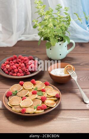 Pancake cereali su una forchetta con yogurt. Lamponi freschi maturi rossi in un piatto marrone e un mazzo di Melissa in una tazza sul tavolo di legno scuro vicino alla W. Foto Stock