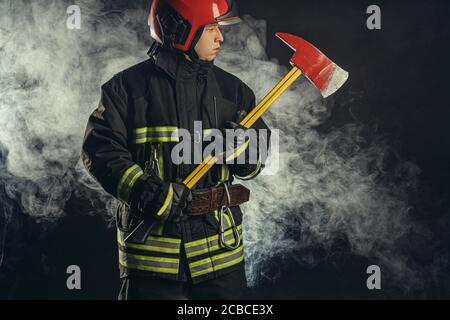 giovane vigile del fuoco che si prepara a salvare, spegnere il fuoco e salvare le persone, se necessario, tenendo il martello in mano e indossando tuta protettiva uniforme Foto Stock