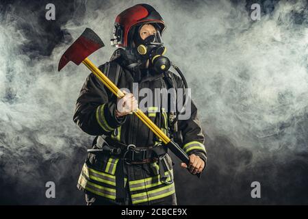 ritratto di serio e sicuro pompiere caucasico regge martello, indossando speciale uniforme protettiva sullo sfondo fumoso Foto Stock