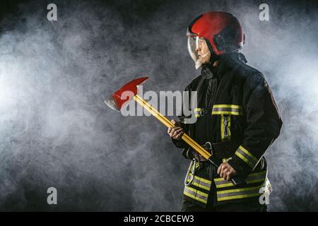 giovane vigile del fuoco che si prepara a salvare, spegnere il fuoco e salvare le persone, se necessario, tenendo il martello in mano e indossando tuta protettiva uniforme Foto Stock