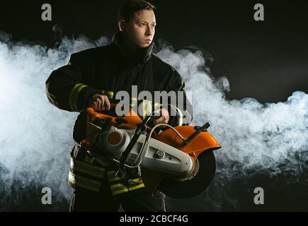 estintore giovane che tiene la motosega stand isolato su sfondo fumoso, indossando un abito da vigile del fuoco, vigile del fuoco sicuro Foto Stock