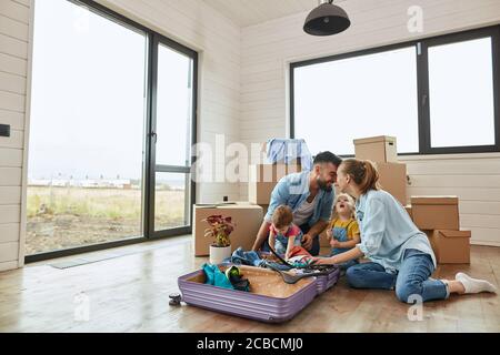 La famiglia caucasica si siede sul pavimento, l'uomo e la donna toccano le fronte, scompattano la valigia in una nuova casa. Dietro di loro spostamento scatole, porta, finestra. Uomo tenere in ginocchio Foto Stock