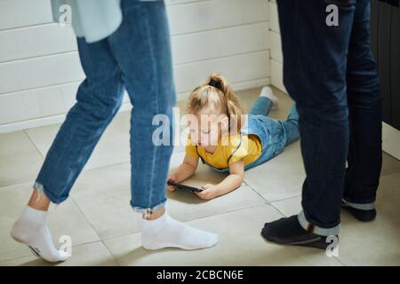 Il bambino caucasico indossa una T-shirt gialla e tute in denim giace sul pavimento e gioca sul telefono. I genitori sono vicini a calze bianche e nere Foto Stock