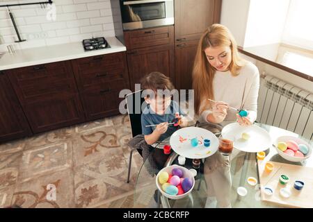 Giovane bella bionda madre e il suo grazioso figlio di quattro anni avendo divertimento mentre la pittura delle uova di Pasqua in cucina domestica, sorridente, sviluppando un creative Foto Stock