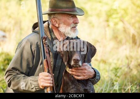 Uomo caucasico maturo con pistola e cane sedersi alla ricerca preda. Uomo bearded in vestiti di caccia. Autunno Foto Stock