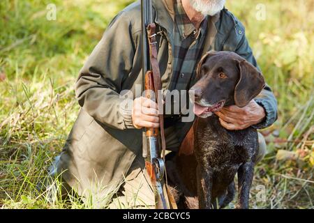 Uomo corto con barba abbraccio il suo cane cacciatore nella foresta d'autunno. Uomo che indossa abiti da caccia casual, seduto su Ground.Gun in mani