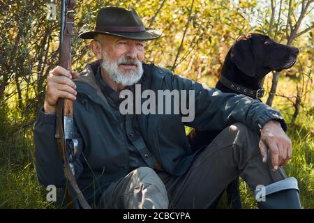 Cane attento e rilassante proprietario seduto su erba della foresta. Uomo in cappello. Che tiene la pistola. Alberi sfondo Foto Stock