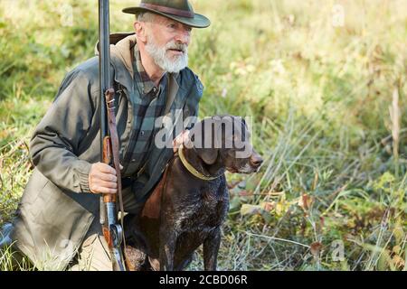 Uomo caucasico maturo con pistola e cane sedersi alla ricerca preda. Uomo bearded in vestiti di caccia. Autunno Foto Stock