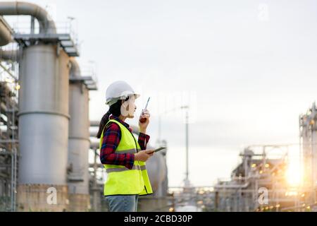 Donna asiatica tecnico industriale ingegnere utilizzando walkie-talkie e tenuta bluprint lavorando in raffineria di petrolio per l'indagine sul sito di costruzione in ingegneria civile Foto Stock