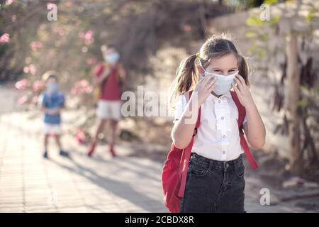 Pupilla con maschera medica sul viso e zaini all'aperto. Educazione durante il periodo del coronavirus. Bambini e assistenza sanitaria. Ritorno a scuola. Foto Stock