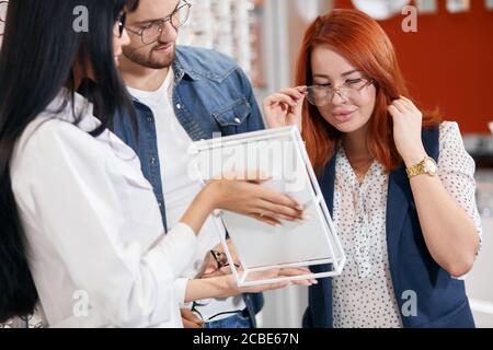 giovane bella donna che indossa occhiali che posano allo specchio, pensando di comprarli o no. zenzero ragazza che prende la decisione befor fare lo sho Foto Stock