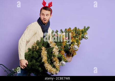 Bel modello maschile in posa su una parete luminosa con albero di Natale splendidamente decorato, indossando divertente archetto rosso, guardando indietro con aperto Foto Stock