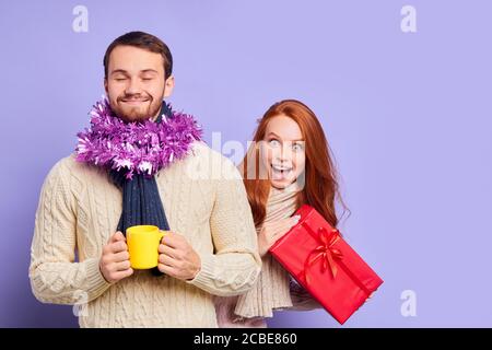 Allegra gioiosa ragazza con regalo di Natale in mano, in piedi dietro un bell'uomo, cercando di fare sorpresa, guardando la macchina fotografica con esprsi sopravorato Foto Stock