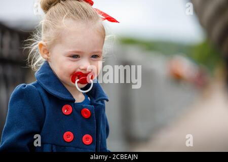 bambina in un cappotto blu con un succhietto dentro la sua bocca sulla strada Foto Stock