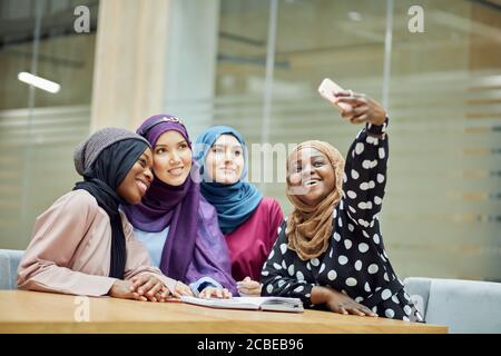 Bella giovane musulmana multietnica gruppo di ragazze a lungo intelligente abiti tradizionali e fiabe che guardano lo smartphone in mano di donna africana mentre s Foto Stock