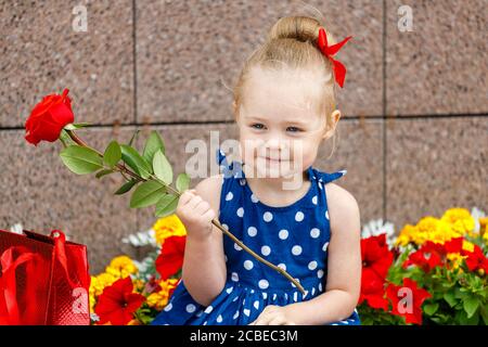 una bambina in un vestito blu e un rosso bow si siede con borse colorate sulla strada accanto a. fiori Foto Stock