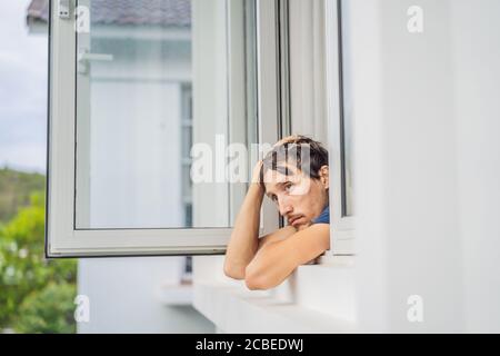 Un giovane estremamente stanco che guarda fuori dalla finestra, casa da sola. Autoisolamento a casa, quarantena a causa della pandemia COVID 19. Problemi di salute mentale in Foto Stock