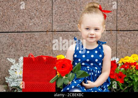 una bambina in un vestito blu e un rosso bow si siede con borse colorate sulla strada accanto a. fiori Foto Stock