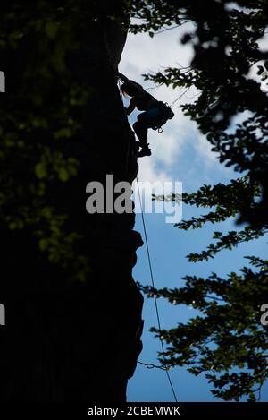 Immagine verticale di una figura femminile silhouette che sale sulla roccia scura contro il cielo blu, foglie verdi sotto. Vista ad angolo basso. Concetto di sport di montagna Foto Stock