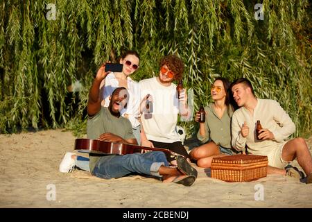 Memoria. Gruppo di amici che indossano bicchieri da birra durante il picnic in spiaggia sotto il sole. Stile di vita, amicizia, divertimento, weekend e riposo concetto. Sembra allegro, felice, festoso, festoso. Foto Stock