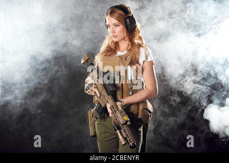Giovane bionda femmina snipper in outfit militare con fucile d'assalto in studio su sfondo fumoso scuro. Concetto di donne in servizio militare. Foto Stock
