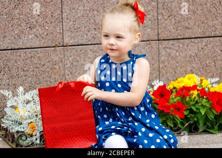 una bambina in un vestito blu e un rosso bow si siede con borse colorate sulla strada accanto a. fiori Foto Stock