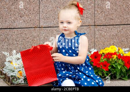 una bambina in un vestito blu e un rosso bow si siede con borse colorate sulla strada accanto a. fiori Foto Stock