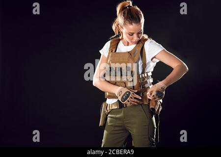 Splendida giovane donna con lunghi capelli biondi che indossa attrezzatura militare, in posa isolata in studio su sfondo scuro Foto Stock