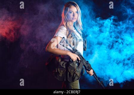 Giovane bionda femmina snipper in outfit militare con fucile d'assalto in studio su sfondo fumoso scuro. Concetto di donne in servizio militare. Foto Stock