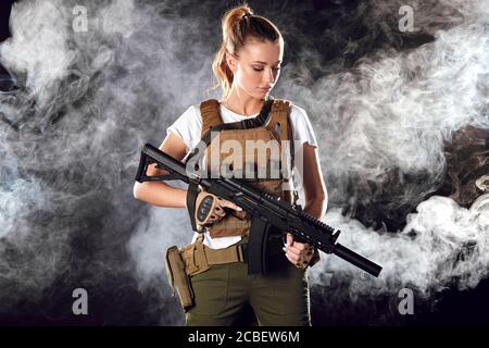 Giovane bionda femmina snipper in outfit militare con fucile d'assalto in studio su sfondo fumoso scuro. Concetto di donne in servizio militare. Foto Stock