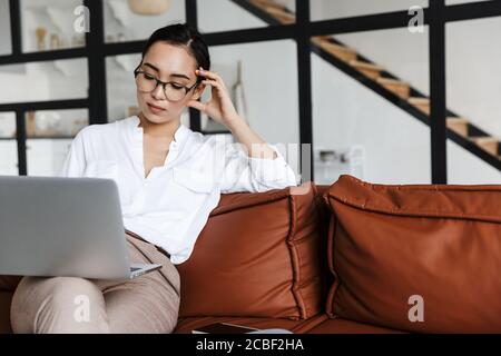 Attraente giovane donna asiatica d'affari che si rilassa su un divano di pelle a casa, lavorando su un computer portatile Foto Stock
