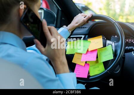 concetto di programma di giorno occupato - donna che guida l'automobile con a. fare un elenco delle note sulla ruota e parlare sul telefono Foto Stock