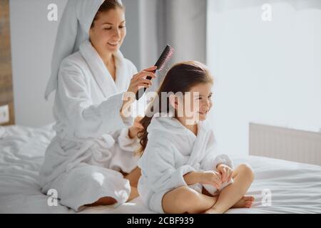 bella donna pettinando i capelli a sua figlia dopo la doccia, seduta sul letto in camera da letto chiara, indossando asciugamano e accappatoio Foto Stock