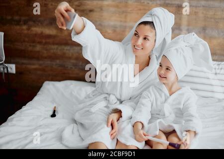 bella donna e bambino che indossa accappatoio e asciugamano bianchi sdraiato sul letto prendendo selfie insieme dopo la doccia Foto Stock