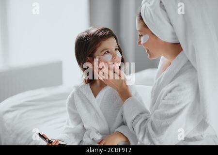 concetto di bellezza. la madre attenta usa le macchie per gli occhi e la figlia in maschera anche. genitori e bambini interessati alle procedure di bellezza Foto Stock