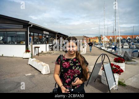 Una ragazza preteen con i capelli marroni lunghi si affaccia la macchina fotografica. Un porticciolo in barca a vela sullo sfondo. Cielo nuvoloso Foto Stock