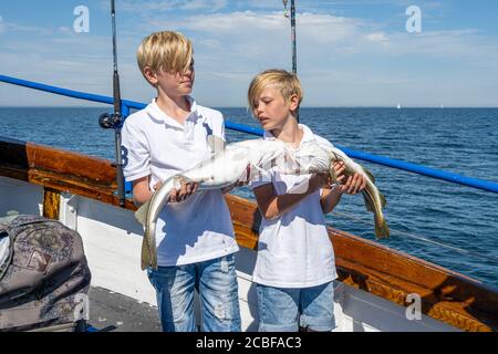 Malmo, Svezia - 6 agosto 2020: Due ragazzi in un viaggio di pesca con la famiglia mostrano la loro cattura fresca di merluzzo. Oceano blu e cielo sullo sfondo Foto Stock