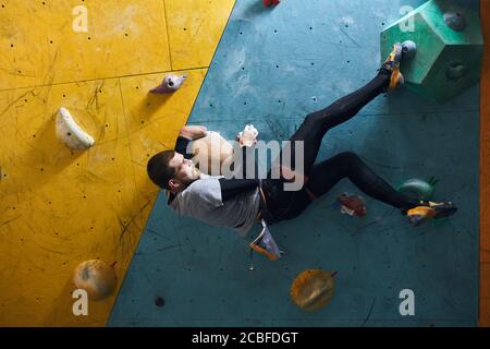 Vista laterale di un masso motivato con disabilità attiva e barba appesa a una parete di arrampicata colorata, che tiene grandi rocce artificiali, cercando di Foto Stock