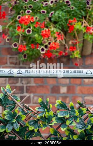 ‘mantenere una distanza di sicurezza di 2 metri’ di nastro di avvertimento davanti a una parete in mattoni con cestello sospeso contenente fiori estivi. Inghilterra Regno Unito Foto Stock